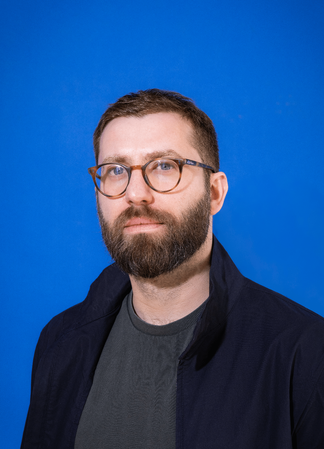 Portrait of a man with brown hair and glasses on a flat gray background.